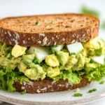 Avocado Egg Salad Sandwich On A White Plate With Fresh Chives And Lettuce, Set Against A Neutral Background
