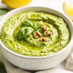 Bowl Of Creamy Avocado Pesto Sauce Garnished With Basil, Olive Oil Drizzle, And Walnuts, With Lemon Wedges On A White Plate