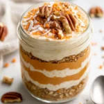 Layered Carrot Cake Overnight Oats In A Glass Jar Topped With Coconut, Pecans, Grated Carrot, And Cinnamon On A White Surface