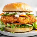 Crispy Cod Sandwich On A White Plate, Topped With Mayo-Pickle Sauce And Fresh Veggies