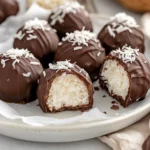 Glossy Homemade Coconut Bonbons On White Plate, Some Garnished With Shredded Coconut, Halved Bonbons Show Creamy Filling.