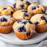 Close-Up Of Keto Blueberry Muffins On A White Plate, Garnished With Powdered Sweetener And Fresh Blueberries