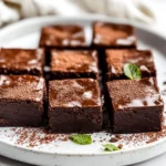 Decadent No-Bake Vegan Brownies Served On A White Plate, Garnished With Cocoa Powder And Fresh Mint Leaves