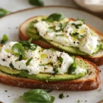 Avocado Toast With Burrata Topped With Fresh Herbs And Aleppo Pepper On A White Plate