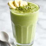 Creamy Banana Matcha Smoothie In A Tall Glass, Garnished With Banana Slices And Matcha Powder