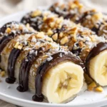 Banana Sushi With Dates On A White Plate, Topped With Coconut And Nuts, Drizzled With Dark Chocolate.