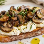 Burrata Toast With Sautéed Mushrooms On A White Plate, Topped With Fresh Rosemary And Drizzled With Olive Oil