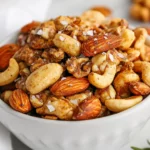 Bowl Of Chili Rosemary Roasted Nuts Garnished With Fresh Rosemary And Flaky Salt On A Neutral Background
