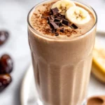 Cold Brew Coffee Smoothie Topped With Chocolate Shavings And Cinnamon, Served In A Tall Glass With A Wooden Spoon Beside It.