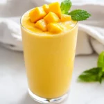 Creamy Mango Smoothie With Almond Milk In A Clear Glass, Garnished With Mango Cubes And Mint Leaves