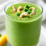 Tropical Green Smoothie In A Glass, Garnished With Lime Zest And Cashews On A White Plate