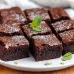 Fudgy Vegan Zucchini Brownies Cut Into Squares On A White Plate, Garnished With Powdered Sugar And Mint Leaves