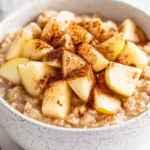 Bowl Of Apple Cinnamon Oatmeal Topped With Apple Slices, Cinnamon, And Maple Syrup On A White Background