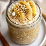 Glass Jar Of Apple Pie Overnight Oats Topped With Yogurt, Cinnamon Apples, Hemp Seeds On A White Plate