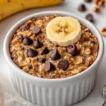 Dish Of Banana Bread Baked Oats In A White Ramekin Topped With Mini Chocolate Chips And Fresh Banana Slices