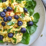 Close-Up Of A Blueberry Breakfast Salad On A White Plate, Featuring Scrambled Eggs, Blueberries, And Fresh Herbs.