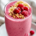 Cranberry Smoothie In A Clear Glass Topped With Raspberries And Banana Slices, Served On A White Plate