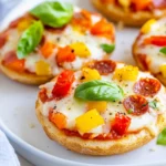 Delicious English Muffin Pizza Topped With Melted Cheese And Colorful Veggies, Served On A White Plate.