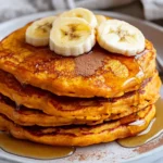 Stack Of Fluffy Sweet Potato Pancakes On A White Plate, Drizzled With Maple Syrup And Garnished With Banana Slices