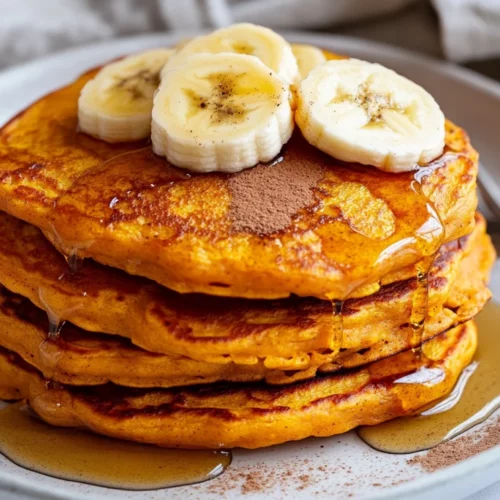 Stack Of Fluffy Sweet Potato Pancakes On A White Plate, Drizzled With Maple Syrup And Garnished With Banana Slices