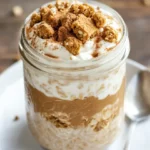 Mason Jar Of Gingerbread Overnight Oats Topped With Greek Yogurt And Gingerbread Cookie Crumbs On A White Plate.