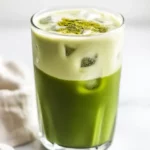 Iced Matcha Latte In A Tall Glass With Ice And Almond Milk, Garnished With Matcha Powder On A White Background