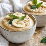 Bowl Of Paleo Banana Pudding Topped With Banana Slices And Coconut Flakes, On A Wooden Table With Mint Leaves