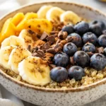 Bowl Of Quinoa Breakfast Bowl Topped With Fresh Fruits, Cacao Nibs, And Maple Syrup On A Wooden Surface