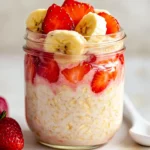 Mason Jar Of Strawberry Banana Overnight Oats Layered With Fresh Fruit And Honey Drizzle On A White Background