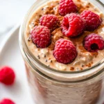 Jar Of Vanilla Latte Overnight Oats Topped With Raspberries And Almond Butter, Served On A White Plate.