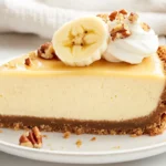 Slice Of Banana Cheesecake On A White Plate, Topped With Fresh Banana Slices And Crushed Pecans