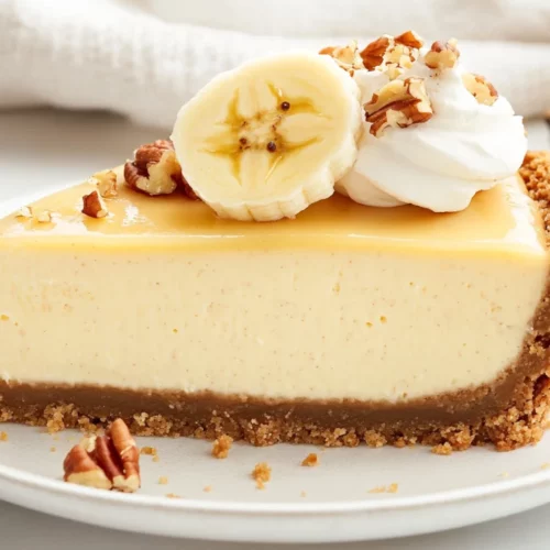 Slice Of Banana Cheesecake On A White Plate, Topped With Fresh Banana Slices And Crushed Pecans