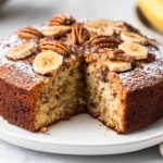 Slice Of Banana Coffee Cake On A White Plate, Topped With Pecans And Powdered Sugar, Showcasing Moist Texture.