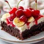 Decadent Banana Split Brownies Topped With Cream Cheese, Cherries, And Banana Slices On A White Plate.