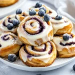 Stack Of Blueberry Cinnamon Rolls Drizzled With Cream Cheese Frosting On A White Plate, Garnished With Blueberries