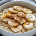 Bowl Of Brown Sugar Banana Oatmeal Topped With Sliced Bananas And Cinnamon On A Neutral Table