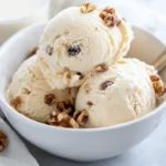Bowl Of Chunky Banana Ice Cream Topped With Walnuts And Raisins, Drizzled With Almond Milk On A Neutral Background.