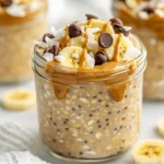 Glass Jar Of Chunky Monkey Overnight Oats Topped With Banana Slices And Cacao Nibs On A White Plate