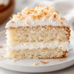 Slice Of Coconut Cream Cake On A White Plate Topped With Whipped Cream And Toasted Coconut Shreds