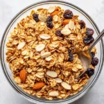Bowl Of Coconut Oil Granola With Almonds, Pistachios, And Golden Raisins Surrounded By A Gray Kitchen Towel