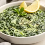 Creamed Spinach Side Dish In A White Bowl Garnished With Black Pepper And Lemon Wedges