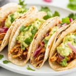 Crispy Crockpot Chicken Tinga Tacos Served On A White Plate, Topped With Avocado Crema And Fresh Herbs