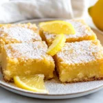 Plate Of Easy Lemon Brownies Topped With Lemon Glaze And Zest, Served On A White Plate With Fresh Lemon Slices.