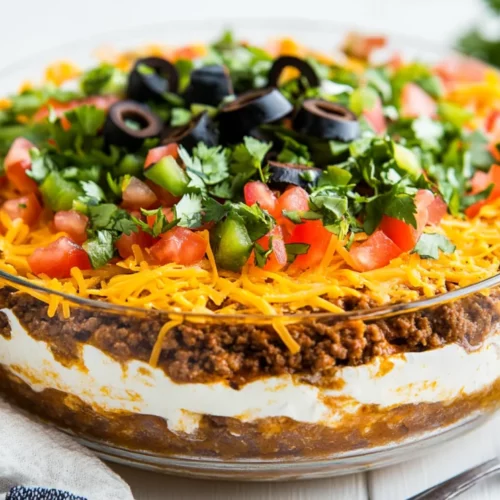 Layered Ground Beef Taco Dip Topped With Lettuce, Cheese, And Tomatoes In A Clear Dish On A White Table