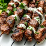 Grilled Lamb Skewers With Mint Sauce On A White Plate, Garnished With Fresh Mint And Lime Wedges.