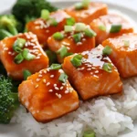 Delicious Orange Salmon Bites On White Plate With Rice And Broccoli, Garnished With Green Onions And Sesame Seeds