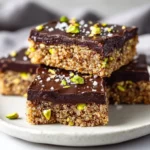 Delicious Salted Dark Chocolate Pistachio Quinoa Bars On A White Plate, Garnished With Chopped Pistachios And Sea Salt