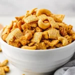 Savory Snack Mix With Cashews In A White Bowl, Showcasing Crispy Cereals And Nuts On A Neutral Table