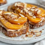 Almond Butter Toast With Peaches, Cinnamon, Toasted Coconut, And Slivered Almonds On A White Plate