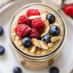 Jar Of Almond Milk Overnight Oats Topped With Fresh Berries And Sliced Almonds On A White Plate